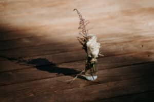 Eine kleine Glasvase enthält zwei weiße Rosen und zarte rosa Zweige und wirft einen sanften Schatten auf eine in warmes, natürliches Licht getauchte Holzoberfläche – ein perfekter Mittelpunkt für eine intime Hochzeitsatmosphäre.