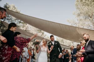 Ein glückliches Paar schreitet im Freien unter einem Baldachin zum Altar, umgeben von einer jubelnden Menge. Die Braut hält einen Blumenstrauß, während die Gäste Blütenblätter werfen. Sie feiern ihre Hochzeit unter einem sonnigen Himmel mit Bäumen im Hintergrund. Es ist der perfekte Höhepunkt einer reibungslosen Hochzeitsplanung.