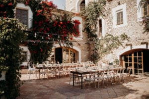 Ein rustikaler Innenhof, perfekt für eine Hochzeit, mit langen Holztischen und Stühlen, umgeben von Steingebäuden. Rote Blumen und Grünpflanzen schmücken die Wände, während darüber Lichterketten funkeln. Die Szene ist in warmes, natürliches Licht getaucht und schafft eine idyllische Kulisse für Ihre Hochzeitsplanung.