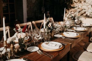 Ein wunderschön gedeckter rustikaler Holztisch, bereit für eine formelle Hochzeitsfeier. Der Tisch ist mit weißen Tellern, elegantem Glasgeschirr, Blumengestecken mit Rosen und großen weißen Kerzen ausgestattet, die eine warme und einladende Atmosphäre schaffen.