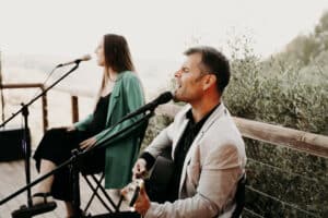 Ein Mann und eine Frau treten im Freien mit Mikrofonen auf, möglicherweise als Unterhaltungskünstler bei einer Hochzeit. Der Mann in einer leichten Jacke spielt Gitarre, während die Frau in einer grünen Jacke neben ihm sitzt. Sie schmücken ein Holzdeck mit üppigem Grün, das ihr romantisches Duett umrahmt.