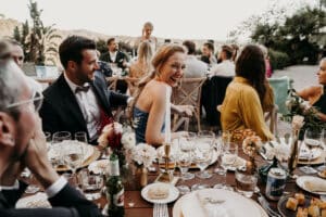 Eine fröhliche Zusammenkunft im Freien zur Feier einer Hochzeit. Die Menschen sitzen formell gekleidet um einen Holztisch, der mit Kerzen, Blumen und Getränken geschmückt ist. Lachen und Gespräche erfüllen die Szene vor einer Kulisse aus Hügeln und Grün.
