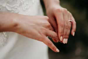 Nahaufnahme einer Person, die einen dünnen Goldring an ihrer linken Hand zurechtrückt und über ihre bevorstehende *Hochzeit* nachdenkt. Die Person hat manikürte Nägel und ihre rechte Hand ruht neben ihrer linken. Der Hintergrund ist verschwommen und hebt ein weißes Kleidungsstück mit Spitzendetails hervor.