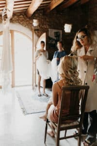 Eine Frau sitzt auf einem Stuhl und lässt sich in einem warm beleuchteten Raum die Haare frisieren, während sie sich auf die Hochzeit vorbereitet. Zwei Personen stehen an einem großen Fenster und halten ein Kleid für die Hochzeit. Neben dem Fenster hängt ein langes weißes Kleid, das sich von rustikalen Holzbalken und Steinwänden abhebt.