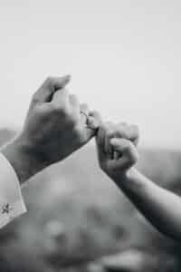 Ein Schwarzweißfoto zeigt zwei Menschen, die ihre kleinen Finger in einer Geste des Versprechens ineinander verschränken und so die Essenz des Heiratens heraufbeschwören. Der sanft verschwommene Hintergrund konzentriert sich auf die Hände, während eine Person ein Hemd mit einem sternförmigen Manschettenknopf trägt, was diesem Moment der Verbundenheit eine elegante Note verleiht.