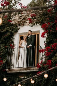 Ein Paar steht auf einem mit roten Blumen und Grünpflanzen geschmückten Balkon und ist ganz in den Moment seiner Hochzeit versunken. Die Frau im weißen Kleid und der Mann im dunklen Anzug blicken in die Ferne, umgeben von einem rustikalen Steingebäude und sanft leuchtenden Lichterketten.