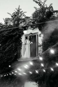 Ein Schwarzweißfoto zeigt Braut und Bräutigam auf einem weinbewachsenen Balkon, wie sie in die Ferne blicken. Lichterketten verschwimmen sanft im Vordergrund und verleihen diesem zeitlosen Moment ihrer Heiratsreise eine romantische Hochzeitsatmosphäre.