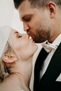 Das Paar, in einen zärtlichen Kuss versunken, fängt die Essenz des Hochzeitsglücks ein. Der Mann trägt einen schwarzen Anzug mit weißer Fliege, während der Schleier der Frau elegant über ihr blondes Haar fällt. Ihre Augen sind geschlossen und sie genießen diesen romantischen Moment, der die Freude des Heiratens ausmacht, innig.