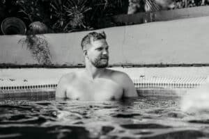 Ein Mann mit Bart entspannt sich in einem Whirlpool, umgeben von einem gefliesten Rand und Pflanzen im Hintergrund. Das in Schwarzweiß aufgenommene Bild ruft die Gelassenheit hervor, die man nach dem Trubel der Hochzeitsplanung sucht.