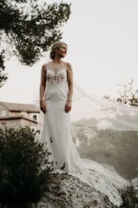 Eine Frau in einem weißen Spitzenbrautkleid steht anmutig auf einem großen Felsen und verkörpert die Essenz einer perfekten Hochzeit. Ihr langer Schleier weht hinter ihr her als Teil dieser wunderschön geplanten Hochzeitsplanung. Im Hintergrund sind Bäume und ein rustikales Gebäude unter einem bewölkten Himmel zu sehen.