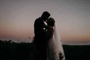 Silhouette einer Braut und eines Bräutigams, die sich bei Sonnenuntergang umarmen, während der Himmel in Rosa- und Lilatönen erstrahlt. Während sie die Details ihrer Hochzeit festlegen, weht der Schleier der Braut sanft in der natürlichen Umgebung, umgeben von üppigem Grün.