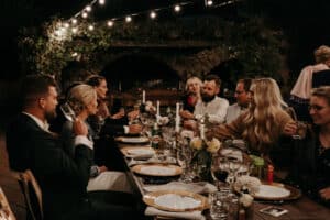 Eine Gruppe von Menschen sitzt im Freien um einen langen Holztisch und feiert ein Hochzeitsessen. Sie sind formell gekleidet, umgeben von Lichterketten und mit Tellern, Gläsern und Blumendekorationen geschmückt. Es ist ein bezauberndes Abendtreffen unter den Sternen.
