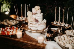 Auf einem rustikalen Hochzeits-Desserttisch steht eine zweistöckige, mit Blumen geschmückte Naked Cake. Um sie herum stehen Cake Pops, Kekse, Obstspieße, Kerzen und Gläser mit Leckereien auf einem Holztisch – die perfekte Note für alle, die gerade mit der Hochzeitsplanung beschäftigt sind.