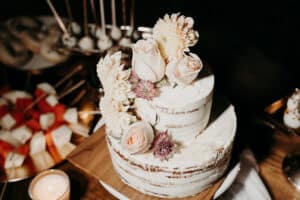 Eine rustikale zweistöckige Torte, perfekt für jede Hochzeit, ist mit weißem Zuckerguss verziert und mit blassrosa Rosen und Chrysanthemen dekoriert. Sie steht auf einem Holzbrett, umgeben von Dessertspießen und einer brennenden Kerze und fängt den zeitlosen Charme einer Hochzeit ein.
