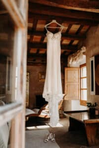 Ein weißes Spitzenbrautkleid hängt elegant an einem Holzbalken in einem rustikalen Raum mit erdfarbenen Wänden und Holzmöbeln und verkörpert die Essenz der Hochzeitsplanung. Sonnenlicht strömt durch offene Fenster herein, während ein Paar weiße High Heels anmutig auf dem Boden darunter ruht, bereit für das freudige Ereignis der Hochzeit.