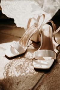 Ein Paar offene Sandalen mit hohem Absatz auf einem Boden mit Spitzenmuster, perfekt für eine Hochzeit. Die beigen Schuhe haben einen Knöchelriemen mit Knopfverschluss. Sonnenlicht wirft weiche Schatten und betont die Spitzenstruktur und die hochzeitsreife Eleganz des Designs.