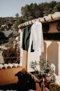 Eine grüne Jacke und ein weißes Hemd hängen an einer rustikalen Steinmauer auf der Terrasse und lassen auf eine elegante Hochzeit unter klarem Himmel schließen. Darunter ruhen braune Schuhe auf einem Holzdeck, umgeben von Terrakotta-Töpfen mit üppigen Pflanzen, während Hügel und Bäume die perfekte Kulisse für Hochzeiten bilden.