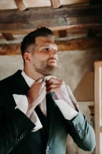 Ein Mann im Anzug richtet seinen Kragen, während er in einem warm beleuchteten Raum mit Holzbalken zur Seite blickt und über seine bevorstehende Hochzeit nachdenkt.
