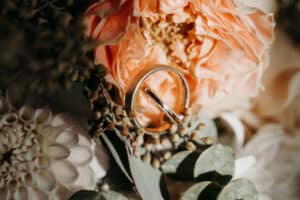 Eine Nahaufnahme von zwei Eheringen, eingebettet zwischen zarten, pfirsichfarbenen Blumen und Grünpflanzen – ein perfektes Herzstück für jede Hochzeit. Das aufwendige Blumenarrangement besteht aus verschiedenen Blüten und Blättern und schafft so ein romantisches und elegantes Bild.