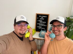 Zwei Menschen, die fröhlich passende Mützen tragen, lächeln, während sie Vintage-Telefone halten – eines gelb und eines blau. Hinter ihnen gibt ein Schild auf Deutsch die Schritte zum Annehmen eines Anrufs an, was ihrem einzigartigen Hochzeits-Fotoshooting eine ungewöhnliche Note verleiht. Eine Pflanze und ein Bücherregal vervollständigen die charmante Szene.