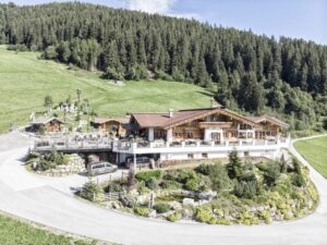 Ein großes Holzhaus, umgeben von üppigen grünen Hügeln und dichtem Wald, unter einem klaren blauen Himmel. Perfekt für eine Traumhochzeit, mit einer großen Außenterrasse mit Sitzgelegenheiten, ideal für Hochzeitsplanung und Feiern, sowie einer kreisförmigen Auffahrt davor.