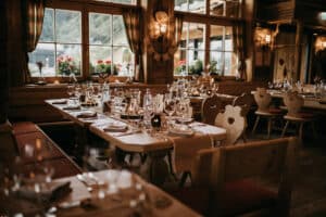 Rustikales Speisezimmer in einem gemütlichen, hölzernen Restaurant mit großen Fenstern, ideal für die Planung einer Hochzeit. Die Tische sind elegant mit Glaswaren, Kerzen und Blumendekorationen arrangiert. Warmes Licht verstärkt die einladende Atmosphäre und die Aussicht auf die Berge bietet eine perfekte Kulisse für Ihren besonderen Tag.