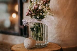 Eine Glasvase mit zarten rosa und weißen Blumen, die mit Spitze zusammengebunden sind, steht auf einer Holzoberfläche und erinnert an die Romantik einer Hochzeit. Daneben befindet sich ein kleines, gesprenkeltes Keramikherz. Ein unscharfer Hintergrund verleiht der Szene eine warme, gemütliche Atmosphäre, perfekt für die Planung Ihres besonderen Tages.