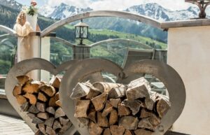 Herzförmige Metallgestelle voller Brennholz schmücken die Terrasse und eignen sich ideal für eine romantische Hochzeitsatmosphäre. Im Hintergrund erheben sich schneebedeckte Berge unter einem wolkigen Himmel, während Glasgeländer den Ausblick einrahmen. Eine Blumenvase ziert das Geländer und verleiht diesem intimen Raum eine elegante Note.