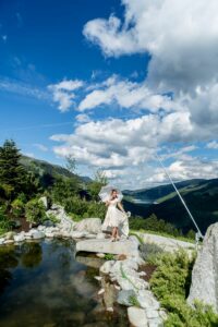 Eine Frau in einem weißen Kleid, die davon träumt zu heiraten, hält einen Sonnenschirm und steht auf Felsen in der Nähe eines reflektierenden Teichs. Die Szene spielt vor einer Kulisse aus sanften grünen Hügeln und einem teilweise bewölkten blauen Himmel.