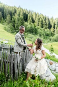 Ein Paar in Hochzeitskleidung posiert auf einem rustikalen Holzzaun inmitten einer üppigen, grünen Landschaft. Die Braut sitzt anmutig mit ihrem Brautstrauß da und umarmt ihren Hochzeitstraum, während der Bräutigam steht und seine Hand reicht. Hohe Bäume und ein strahlend blauer Himmel rahmen diese idyllische Hochzeitsszene ein.