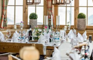Elegantes Speisezimmer für eine Hochzeit in einem rustikalen Raum mit Holztischen, gedeckt mit weißen Tischdecken, Weinflaschen, Gläsern und gefalteten Servietten. Große Fenster und grüne Topfpflanzen sorgen für Charme, während dekorative Kerzen und Blumengestecke das Ambiente bereichern. Perfekt für die Hochzeitsplanung.