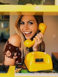 Eine Frau mit langen Haaren lächelt, während sie ein gelbes Retro-Telefon hält und vielleicht ihre Hochzeitsplanung bespricht. Sie trägt ein geblümtes schulterfreies Oberteil und steht an einer leuchtend gelben Theke, im Hintergrund sind Regale voller Bücher und Dekorationen zu sehen.
