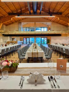 Ein wunderschön eingerichteter Bankettsaal, perfekt für eine Hochzeit, mit langen Tischen, geschmückt mit weißen Tischdecken, elegantem Geschirr, Blumengestecken und Menüs. Holzdecken und moderne Beleuchtung unterstreichen die warme, einladende Atmosphäre, die ideal für eine exquisite Hochzeitsplanung ist.