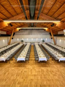 Ein geräumiger Bankettsaal, der sich perfekt für Hochzeitsfeiern eignet, mit langen Reihen elegant gedeckter Tische und grauen Stühlen. Die Holzdecke und der Holzboden verleihen dem Veranstaltungsort Wärme. Teller und Gläser sind ordentlich angeordnet, dekorative Pflanzen im Hintergrund verbessern die Atmosphäre.