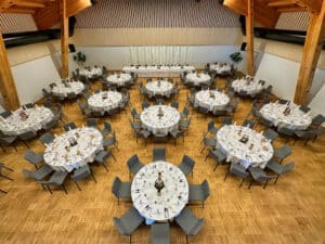 Ein geräumiger Bankettsaal, perfekt für eine Traumhochzeit, mit zehn runden Tischen mit weißen Tischdecken und grauen Stühlen. Der Raum verfügt über Holzbalken und eine hohe Decke, die eine elegante Atmosphäre schaffen, die ideal für Hochzeiten ist. Zwei Topfpflanzen schmücken den Raum.