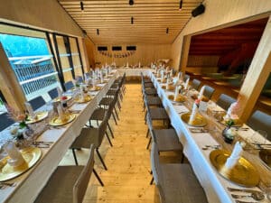 Eleganter Bankettsaal, perfekt für eine Hochzeit, mit zwei langen Tischen, die für eine formelle Veranstaltung gedeckt sind. Jeder Tisch ist mit goldenen Tellern, weißen Servietten und Blumendekorationen geschmückt. Der Raum verfügt über Holzwände und -decken und große Fenster bieten eine malerische Aussicht, ideal für diejenigen, die ihren Traumtag planen.
