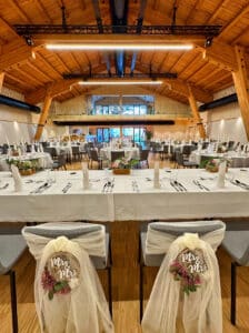 Ein wunderschön dekorierter Hochzeitsort mit gedeckten Tischen für die Gäste. Zwei Stühle im Vordergrund sind mit „Mr & Mrs“-Schildern mit floralen Akzenten versehen, was exquisite Hochzeitsplanung widerspiegelt. Der Raum ist mit einer Holzbalkendecke und großen Fenstern im Hintergrund warm beleuchtet.