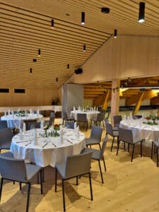 Ein moderner Bankettsaal mit Holzdecke und -boden, gedeckt für eine Hochzeit. Runde, mit weißen Tischdecken bedeckte Tische sind elegant mit Servietten, Gläsern und kleinen Blumengestecken arrangiert. Graue Stühle umgeben die Tische, perfekt für Ihre Hochzeitsplanung.