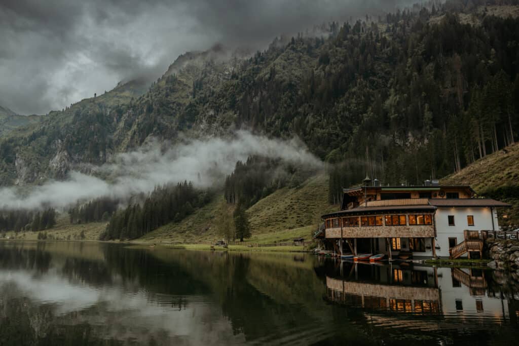 Ein ruhiger See spiegelt eine Holzhütte am Fuße eines nebligen, bewaldeten Berges wider – eine perfekte Kulisse für eine Traumhochzeit. Niedrige Wolken ziehen durch die immergrünen Bäume und schaffen eine stimmungsvolle und ruhige Szene unter einem bewölkten Himmel, ideal für Hochzeiten.