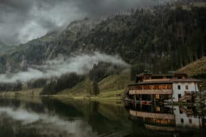 Ein ruhiger See spiegelt eine Holzhütte am Fuße eines nebligen, bewaldeten Berges wider – eine perfekte Kulisse für eine Traumhochzeit. Niedrige Wolken ziehen durch die immergrünen Bäume und schaffen eine stimmungsvolle und ruhige Szene unter einem bewölkten Himmel, ideal für Hochzeiten.