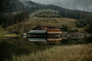 Ein rustikales Gebäude inmitten üppiger Bäume spiegelt sich in einem ruhigen See und bietet die perfekte Kulisse für eine Hochzeit. Der bewölkte Himmel verleiht der ruhigen Landschaft eine stimmungsvolle Atmosphäre, ideal für diejenigen, die inmitten der Natur heiraten möchten. Die umliegenden Wälder und Hügel tragen zur ruhigen Szene bei.