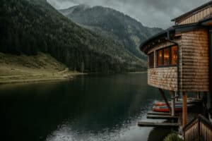 Eine ruhige Holzhütte erstreckt sich über einem ruhigen See mit Bergen und dichten Kiefernwäldern im Hintergrund. Der Himmel ist bedeckt und verbreitet eine heitere, stimmungsvolle Atmosphäre – eine perfekte Kulisse für eine romantische Hochzeit inmitten malerischer Landschaften.