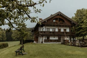 Ein rustikales Holzhaus mit schrägem Dach liegt inmitten üppiger Vegetation, perfekt für eine romantische Hochzeit. Ein Holzzaun und eine Bank im Vordergrund verstärken die ruhige, ländliche Atmosphäre unter bewölktem Himmel, ideal für eine märchenhafte Hochzeitsplanung.