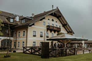 Ein großes, rustikales gelbes Haus mit weißen Zierleisten und einem Holzbalkon bietet die perfekte Kulisse für eine Hochzeit. Der mit einem großen Sonnenschirm geschmückte Sitzbereich im Freien lädt die Gäste zum Feiern inmitten von üppigem grünem Gras und einem romantischen, bewölkten Himmel ein.