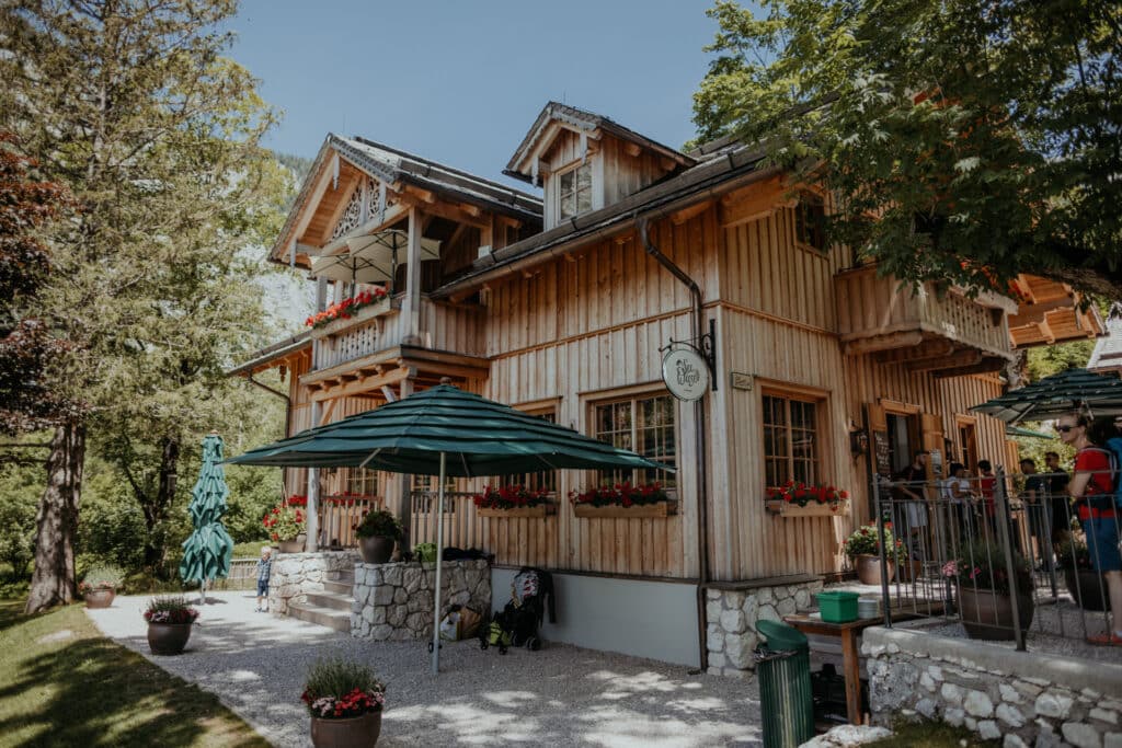 Ein charmantes Holzhaus mit roten Blumenkästen und grünen Sonnenschirmen, perfekt für eine Hochzeit, inmitten üppiger Vegetation und Bäume. Ein kleiner Kiesweg führt zum Eingang. Das Gebäude hat Balkone und im Sitzbereich im Freien sieht man mehrere Personen.