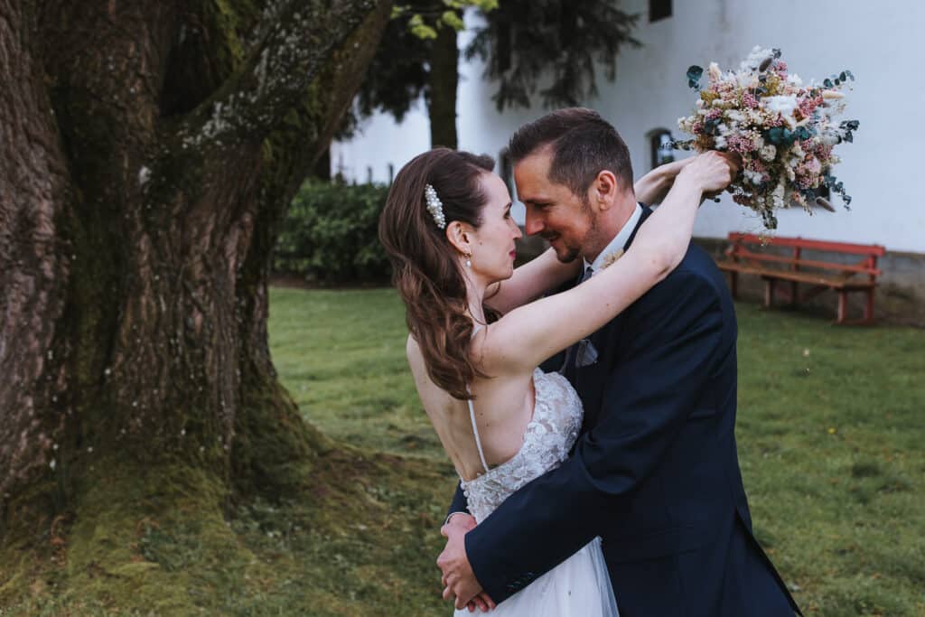 Braut und Bräutigam umarmen sich unter einem großen Baum auf einer Wiese und feiern ihre Hochzeit. Die Braut hält ihren Brautstrauß hoch, während sie sich anlächeln. Im Hintergrund vervollständigen eine Holzbank und ein weißes Gebäude diese malerische Liebesszene.
