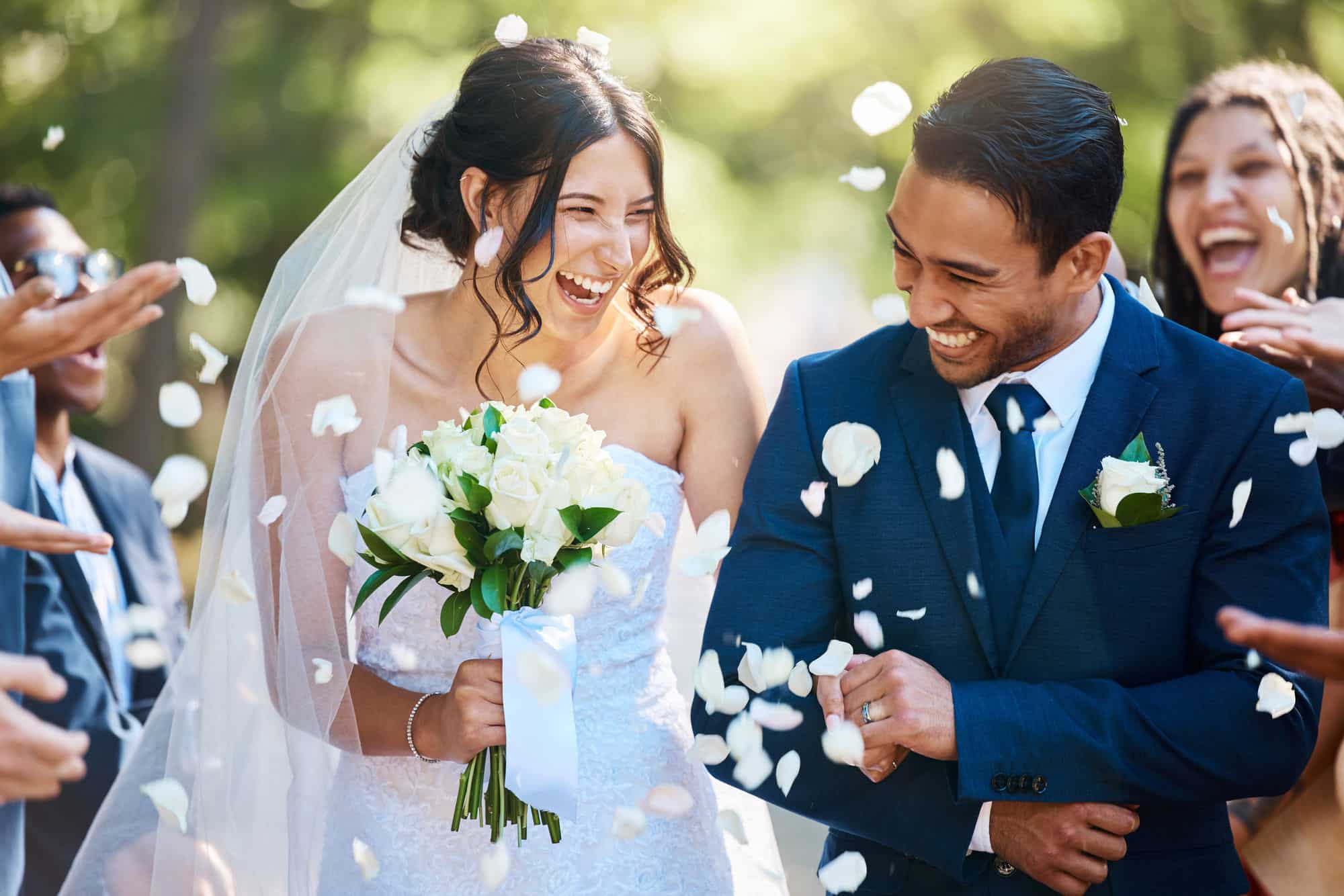 Eine fröhliche Braut und ein Bräutigam im Hochzeitskleid schreiten an ihrem Hochzeitstag im Freien einen Gang entlang, umgeben von Freunden, die Rosenblätter werfen. Die Braut hält einen Strauß weißer Rosen und beide lächeln vor einer sonnigen, grünen Kulisse.