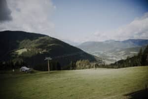 Ein malerischer Blick auf ein üppiges grünes Tal, perfekt für eine Hochzeit, mit Bergen im Hintergrund unter einem teilweise bewölkten Himmel. Eine Seilbahn ziert die linke Seite, während ein Waldgebiet die Ränder des idealen Ortes für Hochzeitsplanung auf einer offenen Wiese säumt.