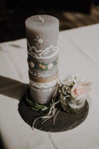 Eine dekorierte Kerze mit Berg- und Blumenmotiven sitzt auf einem Schiefersockel, perfekt für jede Hochzeit. Daneben steht ein kleines Gefäß mit einer rosa Rose und weißen Blumen. Die Kerze trägt die Aufschrift „Lisa & Stefan“ und „25. Juni 2021“ und ist damit eine ideale Ergänzung für Ihre Hochzeitsplanungsdekoration.