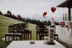 Eine Hochzeit im Freien auf einem grasbewachsenen Hügel mit nach vorne gerichteten Holzstühlen verkörpert die Essenz einer perfekten Hochzeitsplanung. Im Hintergrund schweben rote herzförmige Luftballons und sorgen für eine romantische Note. Auf einem Tisch im Vordergrund stehen eine große dekorierte Kerze und kleine Blumenarrangements für eine unvergessliche Hochzeit.